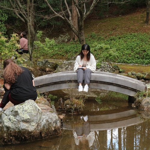 A student sitting on a small bridge overlooking a pond, writing in a journal.